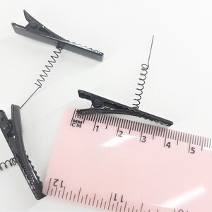 May include: Three black metal hair clips with spring coils. The clips are approximately 4 cm long and are shown next to a ruler.