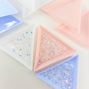 May include: Four triangular trays, each a different colour, filled with clear iridescent glass beads. The trays are white, light blue, pink, and dark blue.
