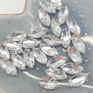 May include: A collection of clear, marquise-shaped rhinestones with multiple facets, reflecting light. The gemstones are scattered on a light blue, textured surface with gold flecks. These rhinestones are suitable for jewelry making and crafts.