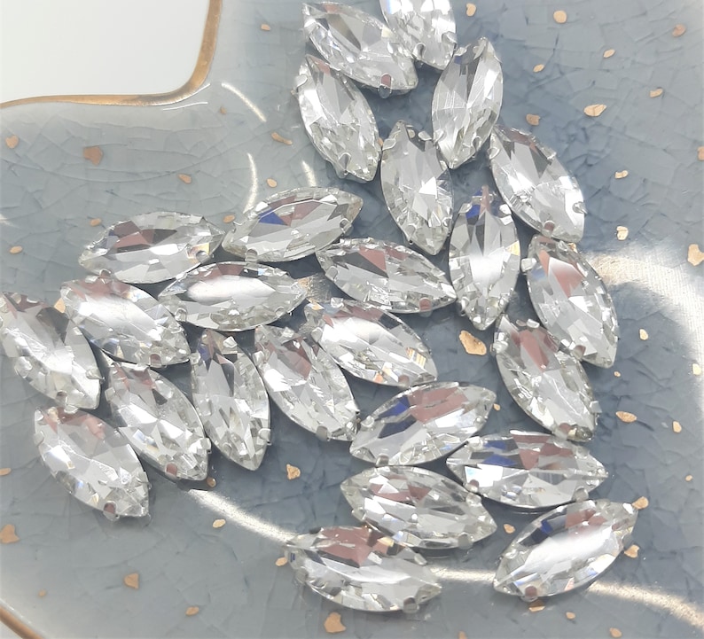 May include: A collection of clear, marquise-shaped rhinestones with multiple facets, reflecting light. The gemstones are scattered on a light blue, textured surface with gold flecks. These rhinestones are suitable for jewelry making and crafts.