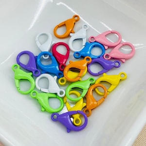 May include: A collection of colorful lobster claw clasps in various shades, including orange, white, pink, blue, purple, green, and yellow. These metal clasps are arranged on a white surface, showcasing their vibrant hues and functional design.