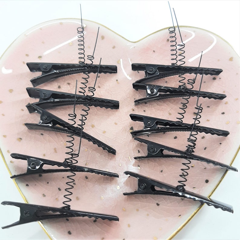 May include: A set of ten black hair clips with a spring coil on each end. The clips are arranged on a pink heart-shaped plate.