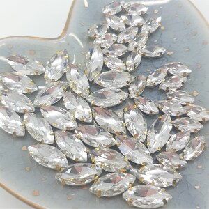 May include: A collection of clear, marquise-shaped rhinestones with gold-colored metal settings. The rhinestones are scattered on a light blue, heart-shaped dish. The gems are faceted, reflecting light and creating a sparkling effect.