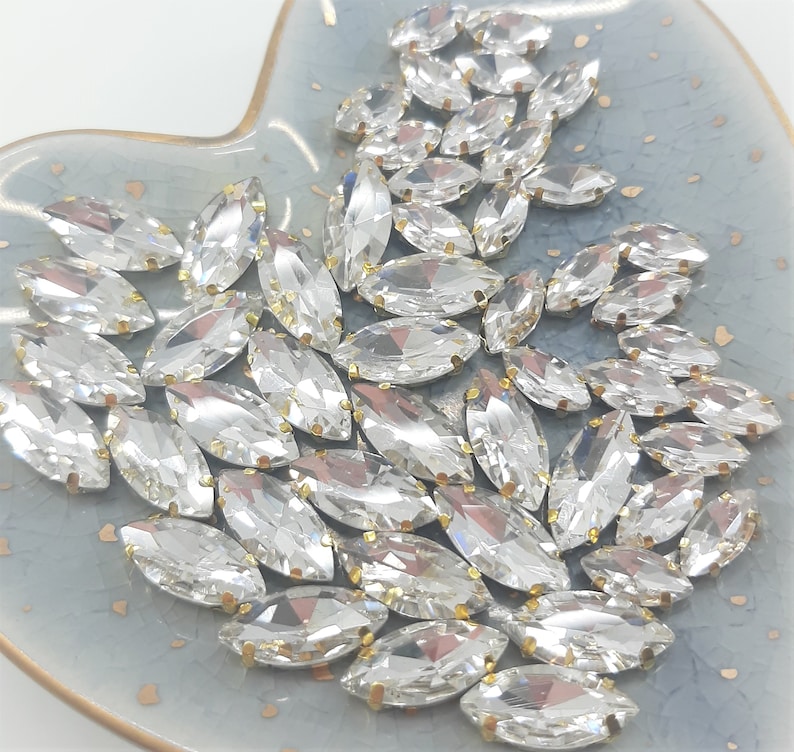 May include: A collection of clear, marquise-shaped rhinestones with gold-colored metal settings. The rhinestones are scattered on a light blue, heart-shaped dish. The gems are faceted, reflecting light and creating a sparkling effect.