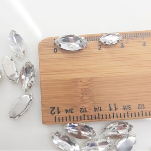 May include: Clear, marquise-shaped rhinestones with silver-colored metal backing are scattered across a white surface. A wooden ruler with inch markings is in the frame, showing the size of the rhinestones. These rhinestones are ideal for embellishing clothing or accessories.