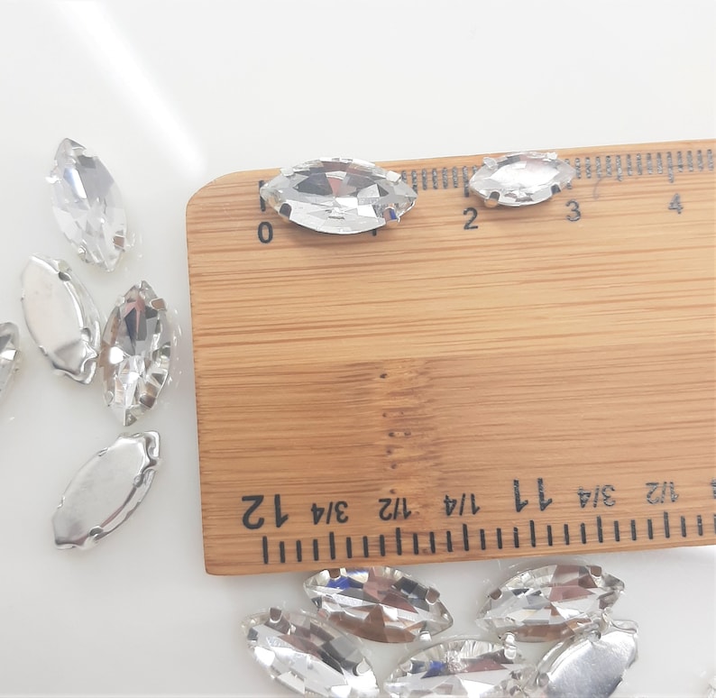 May include: Clear, marquise-shaped rhinestones with silver-colored metal backing are scattered across a white surface. A wooden ruler with inch markings is in the frame, showing the size of the rhinestones. These rhinestones are ideal for embellishing clothing or accessories.