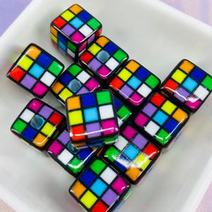 May include: A selection of cube-shaped beads with a vibrant grid pattern in colours such as red, blue, green, yellow, and white. The beads have a glossy finish and are presented in a white square dish.