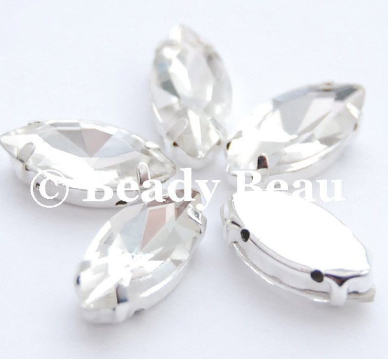 May include: Five clear, marquise-shaped rhinestones with silver-colored metal settings. The rhinestones are faceted, reflecting light. The image includes the text "Beady Beau".