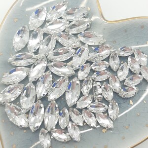 May include: A collection of clear, marquise-shaped rhinestones, displayed on a light blue, heart-shaped dish with a gold rim. The rhinestones are faceted, reflecting light, and are arranged in a scattered pattern.