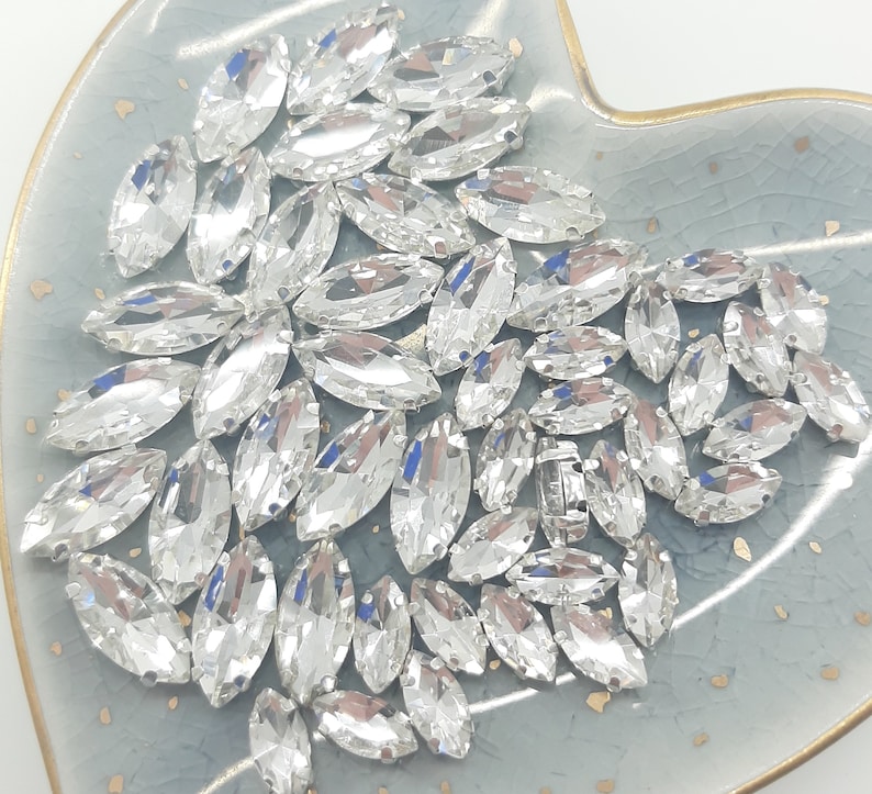 May include: A collection of clear, marquise-shaped rhinestones, displayed on a light blue, heart-shaped dish with a gold rim. The rhinestones are faceted, reflecting light, and are arranged in a scattered pattern.