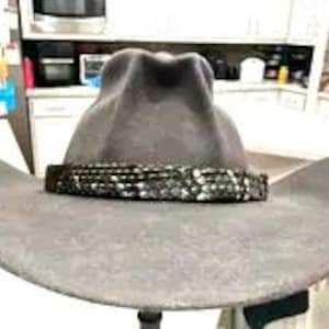 May include: A grey cowboy hat featuring a wide, curved brim and a high, pinched crown. A dark, textured band with a repeating metallic scale-like pattern encircles the base of the crown, providing a distinct contrast to the solid grey felt material.