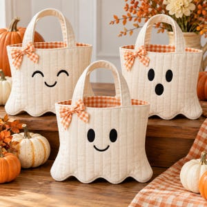 May include: Three off-white quilted fabric tote bags shaped like ghosts with scalloped bottom edges. Each bag features a black embroidered face, two handles, and an orange and white gingham bow. The interior lining is orange and white gingham.