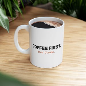 May include: A white ceramic mug filled with black coffee, featuring the black text COFFEE FIRST. and the orange text then Claude. printed on the front. The mug has a curved handle on the left side and a smooth, glossy finish throughout the exterior.