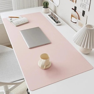 May include: A large, rectangular, light pink desk mat covering a white desk surface. The mat features a silver laptop, a white ceramic mug, a notebook with a pen, and a small electronic device with vertical tubes arranged on its top surface.