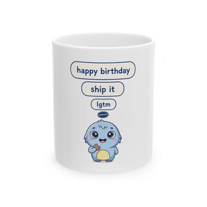 May include: A white ceramic mug featuring a cartoon blue creature holding a cookie. Above the creature are three speech bubbles containing the text "happy birthday", "ship it", and "lgtm", with a small blue "APPROVED" stamp above the creature's head.