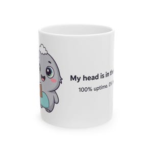 May include: A white ceramic mug featuring a cartoon grey creature with a blue belly and a small white tuft on its head. The creature holds a brown book. The text reads: My head is in the cloud. 100% uptime. 0% focus.