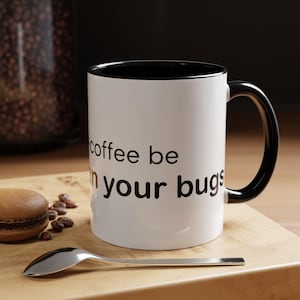 May include: A white ceramic mug with a black interior and handle. The mug features black text that reads "coffee be than your bugs". The mug is positioned on a wooden surface next to a silver spoon and a brown cookie with scattered coffee beans.