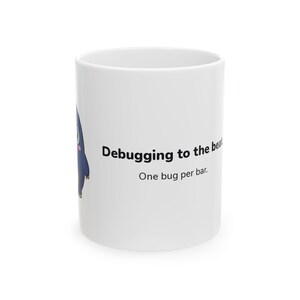 May include: A white ceramic mug featuring a dark blue cartoon character with a yellow belly and the black text "Debugging to the beat. One bug per bar." The mug has a cylindrical shape with a smooth finish and a simple, clean design.