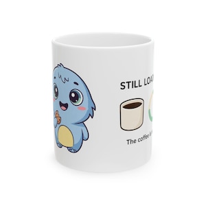 May include: A white ceramic mug featuring a cartoon blue creature with a yellow belly eating a cookie. The text reads STILL LOADING with an illustration of a coffee cup and a partial loading circle, followed by the text The coffee is loading.