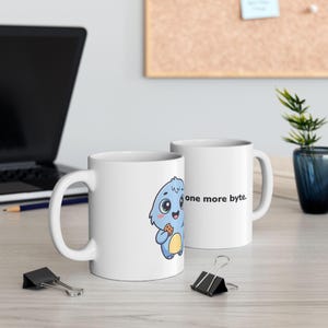 May include: Two white ceramic mugs featuring a cartoon illustration of a light blue creature with a yellow belly holding a small brown cookie. The text "one more byte." is printed in black on the side of the mugs, which have a handle and a cylindrical shape.