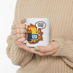 May include: A white ceramic mug featuring a cartoon graphic of a smiling laptop and a server rack engulfed in orange and yellow flames. A speech bubble above the laptop contains the black text: DEPLOYED ON FRIDAY. IT'S FINE. The mug has a handle.