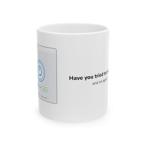 May include: A white ceramic mug featuring a graphic of a power button icon and the text "Have you tried turning it off and on again?" printed in black. The design includes a light grey rectangular box with a blue power symbol and a green button.