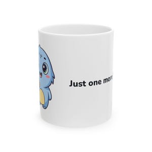 May include: A white ceramic mug featuring a cartoon illustration of a small blue creature with large eyes and a yellow belly. The text "Just one more" is printed in black sans-serif font on the side of the mug, positioned next to the character.