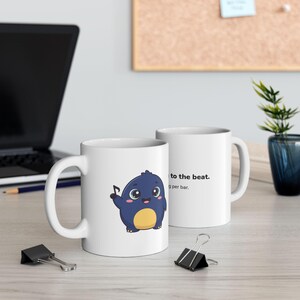May include: Two white ceramic mugs. One mug features a cartoon illustration of a round, dark blue creature with a yellow belly, holding a black musical note. The other mug displays the text: to the beat. 9g per bar.