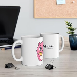 May include: A white ceramic mug featuring a cartoon pink monster holding a taco. The text on the mug reads Build: failed Coffee: passed. The mug has a standard handle and a cylindrical shape, presented as a clean, functional beverage container.