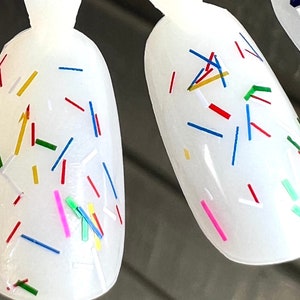 May include: White nail polish with colorful confetti-like glitter. The glitter is in various shapes and sizes, including long, thin strips and small, round pieces. The colors of the glitter include red, blue, green, yellow, and pink.