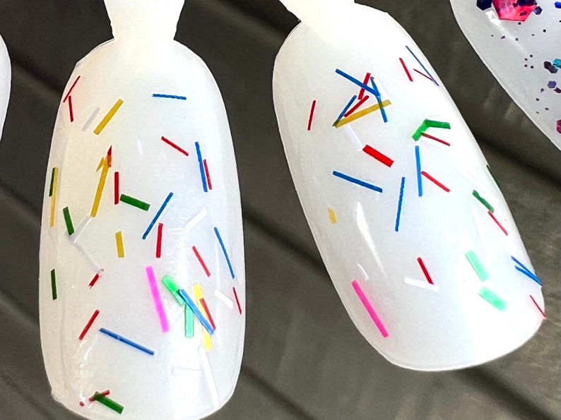 May include: White nail polish with colorful confetti-like glitter. The glitter is in various shapes and sizes, including long, thin strips and small, round pieces. The colors of the glitter include red, blue, green, yellow, and pink.