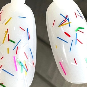 May include: Two white oval-shaped nail polish bottles with a clear, glossy finish. The bottles are decorated with colorful confetti-like sprinkles in red, blue, yellow, green, and pink.