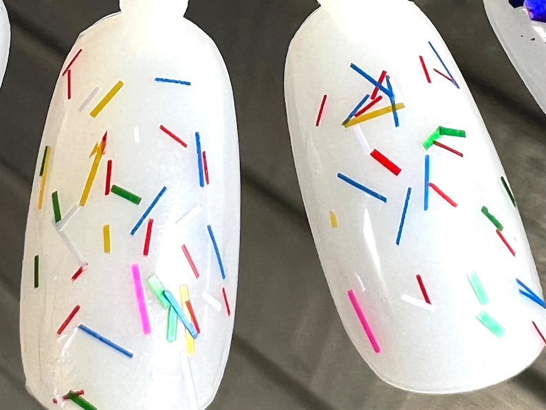 May include: Two white oval-shaped nail polish bottles with a clear, glossy finish. The bottles are decorated with colorful confetti-like sprinkles in red, blue, yellow, green, and pink.