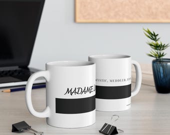 Könnte beinhalten: Zwei weiße Keramikbecher mit einem horizontalen schwarzen Streifen und schwarzem Text. Ein Becher zeigt den Schriftzug "MADAME L", der andere "MYSTIC, MEDDLER, ICON". Beide Becher haben eine zylindrische Form und einen Henkel.