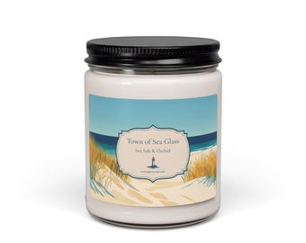 Könnte beinhalten: Eine weiße Kerze in einem klaren Glasgefäß mit schwarzem Deckel. Das Etikett zeigt eine Küstenillustration mit Sanddünen, Strandgras und dem Meer. Der Text lautet Town of Sea Glass und Sea Salt & Orchid mit einem Leuchtturm-Symbol.