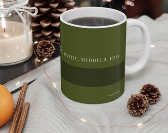Könnte beinhalten: Eine weiße Keramiktasse mit dunkelolivgrüner Außenseite und einem horizontalen Streifen in einem dunkleren Grünton. Der Text "MYSTIC, MEDDLER, ICON" ist in Weiß auf dem oberen Teil der Tasse aufgedruckt, die einen weißen Henkel hat.