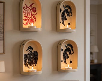 May include: Four arched wall-mounted candle holders made of cream-coloured ceramic. Each features a unique black or red graphic illustration inside the arch, including a pomegranate, a bird, and two celestial-themed designs with stars.