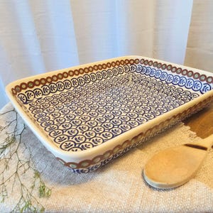 May include: A rectangular ceramic baking dish featuring a repeating blue leaf pattern on a white background, bordered by a decorative band of brown and green circles. The dish has a smooth, glazed finish and a deep interior for food preparation.