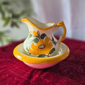 May include: A ceramic creamer pitcher and matching small bowl set featuring a hand-painted yellow flower design with green leaves. Both pieces have a white base and a textured, sponge-painted yellow rim. The pitcher has a curved handle.