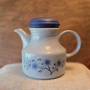May include: A ceramic teapot with a speckled light gray finish and a dark blue lid. The body features a blue floral branch pattern with detailed blossoms and stems. It has a curved handle on the right side and a short spout on the left side.