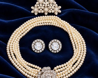 Puede incluir: Un collar de perlas de varias hileras con un broche central de flores con pedrería, un broche triangular a juego con flores de pedrería y dos pendientes redondos con una piedra central grande rodeada de pequeñas piedras.