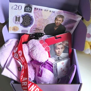 May include: A purple gift box containing a Bank of BTS £20 note, a red lanyard with white text, a purple plush toy with a ribbon, a photo card of a person in a red holder, and a small clear case with the text O! R U L8, 2?