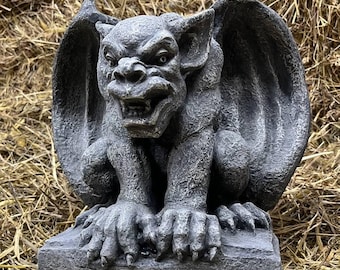 May include: A gray stone gargoyle statue featuring a crouching creature with large, folded wings, pointed ears, and a snarling expression with visible teeth, perched on a rectangular base with a textured, weathered surface finish.