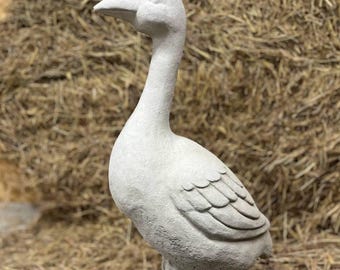 May include: A light gray concrete goose statue standing on a textured base. The sculpture features a long neck, a detailed beak, and carved wing feathers, capturing the bird in a side profile view with its head turned slightly outward.
