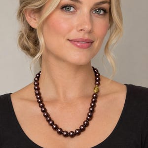 May include: A necklace featuring a single strand of round, dark purple, lustrous beads. The necklace includes a small, textured, gold-toned metal bead positioned asymmetrically on the right side, adding a distinct metallic accent to the dark beads.