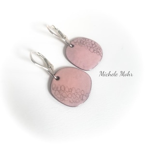 May include: A pair of pink enamel earrings with silver-colored lever-back closures. Each earring features a rounded shape with a textured, dark gray pattern on the top half. The text "Michele Mohr" is visible.