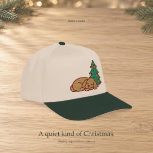 May include: A cream-colored baseball cap with a dark green brim. The front panel features an embroidered graphic of a brown dog sleeping next to a green Christmas tree with orange ornaments. The text ACORN & EMBER and A quiet kind of Christmas.