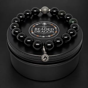 May include: A beaded bracelet featuring smooth, glossy black spherical beads, a single textured silver-tone metal bead, and a small circular silver-tone charm engraved with a dragon silhouette, resting on a black circular tin with the text THE BEADED DRAGON.