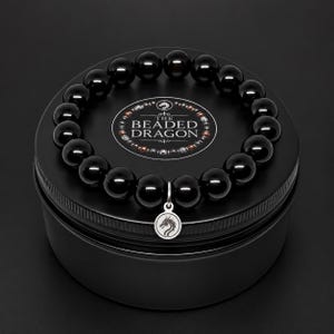 May include: A circular black storage tin topped with a bracelet made of smooth, polished black beads. A silver-toned circular charm featuring a dragon silhouette hangs from the bracelet. The lid displays the text THE BEADED DRAGON.