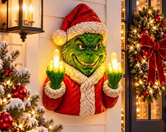 May include: A wall-mounted green character decoration wearing a red Santa hat and coat with white fur trim. The figure has a mischievous expression and is holding up both middle fingers, which are illuminated with a warm yellow light.
